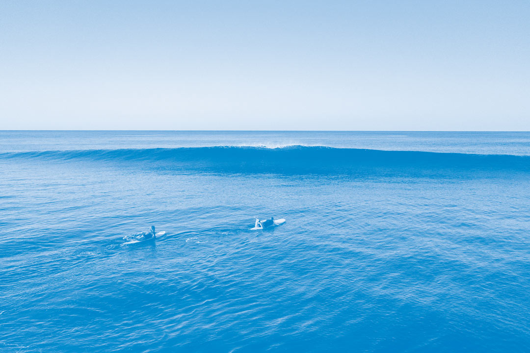 Foto aérea de dos surfistas remando hacia una ola rompiente con una superposición de color azul en la imagen.