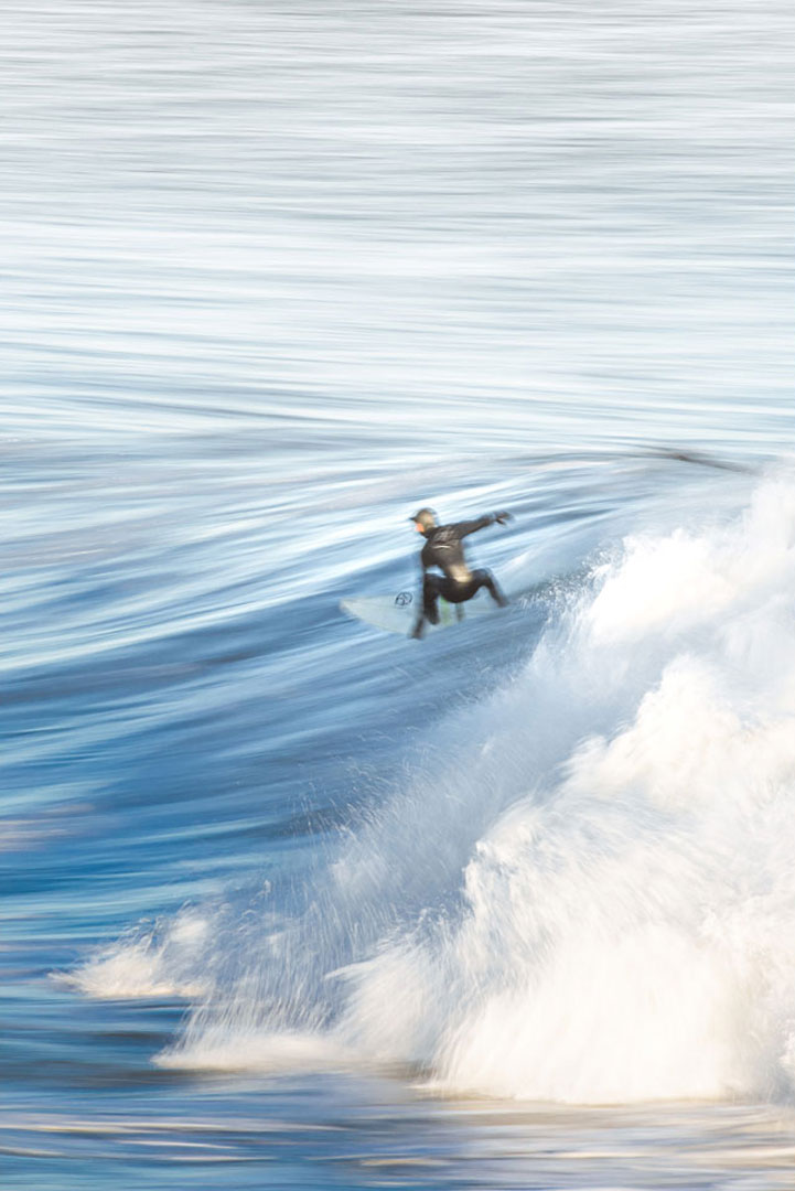 Blurred action shot of a surfer in a wetsuit riding the smooth part of a wave while the white water crashes in the foreground.