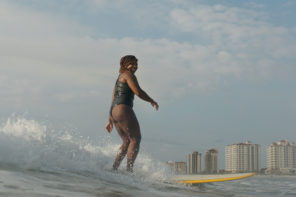 Photo of Gigi Lucas surfing on a yellow surfboard, wearing a black bathing suit, just off the coast.