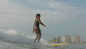 Photo of Gigi Lucas surfing on a yellow surfboard, wearing a black bathing suit, just off the coast.