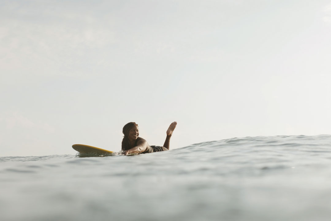 Photo of GiGi Lucas paddling in the ocean on her surfboard, waiting for a wave. The photo is taken from the eye level of the water.