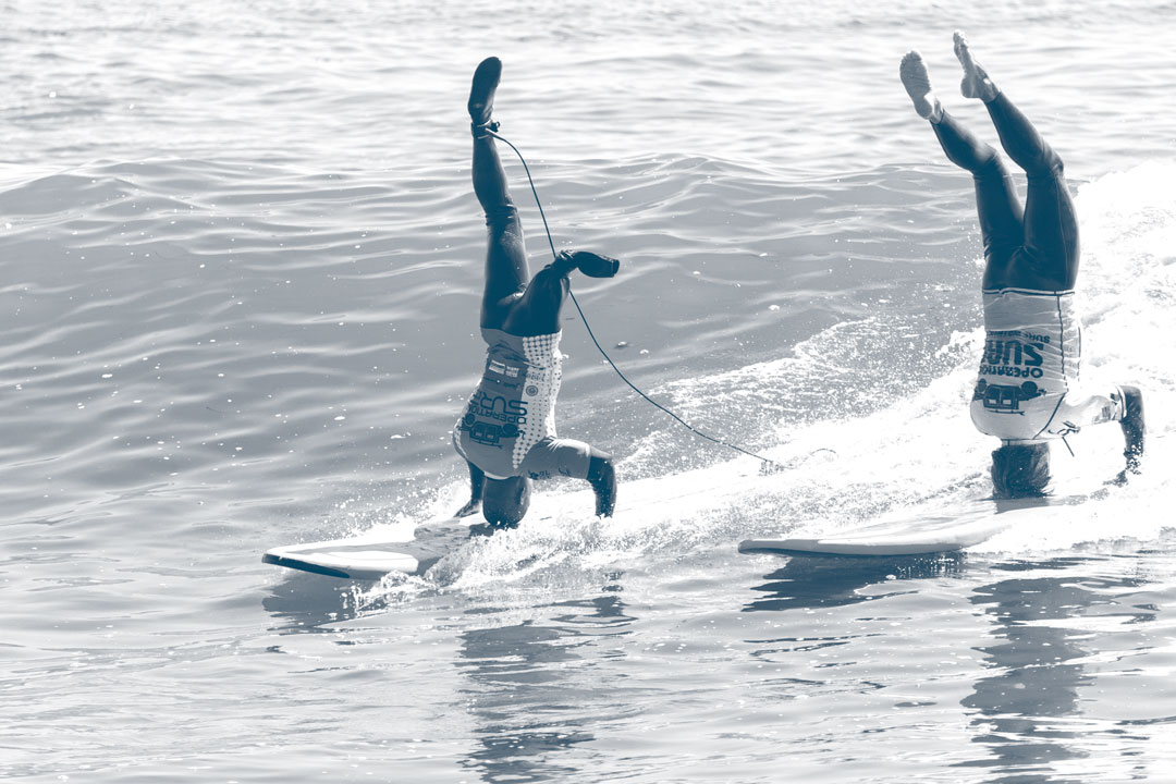 Photo from Operation Surf of two surfers, one with two legs and the other with only one, riding a wave next to each other while doing headstands on their boards.