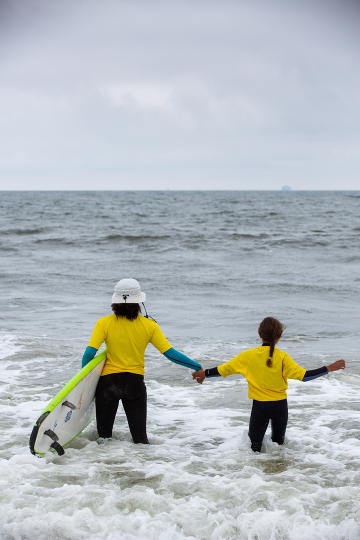 Two girls walking into the ocean together, holding hands, to go surf. The older one is carrying the surfboard while she guides the younger girl out.