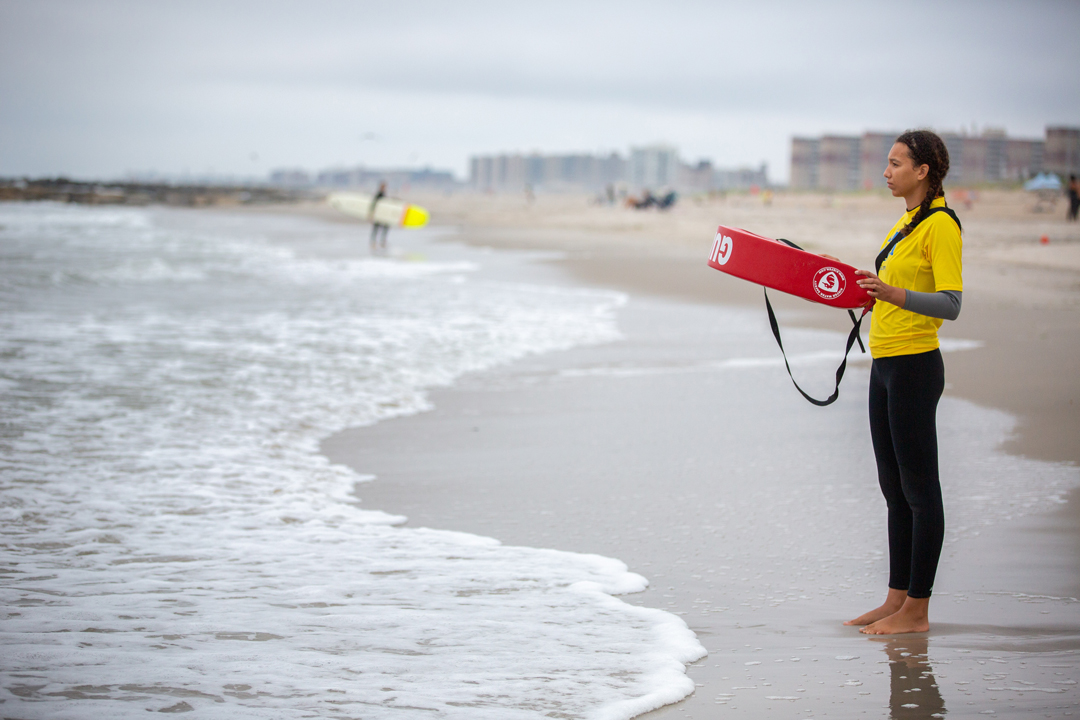 A young girl stands on the beach holding a read life preserver as she watches out in the waves for the surfers.