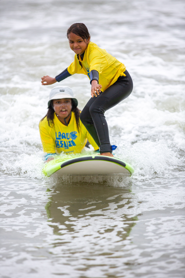 Two girls surfing together. The older girl hangs on to the back of the board while the younger girl is standing on the board surfing.