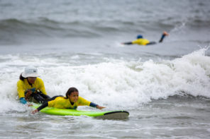 Young girl getting pushed into a wave by her instructor as she paddles on her surfboard. Her tongue is sticked out of her mouth as she focuses on the wave.