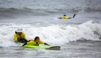 Young girl getting pushed into a wave by her instructor as she paddles on her surfboard. Her tongue is sticked out of her mouth as she focuses on the wave.