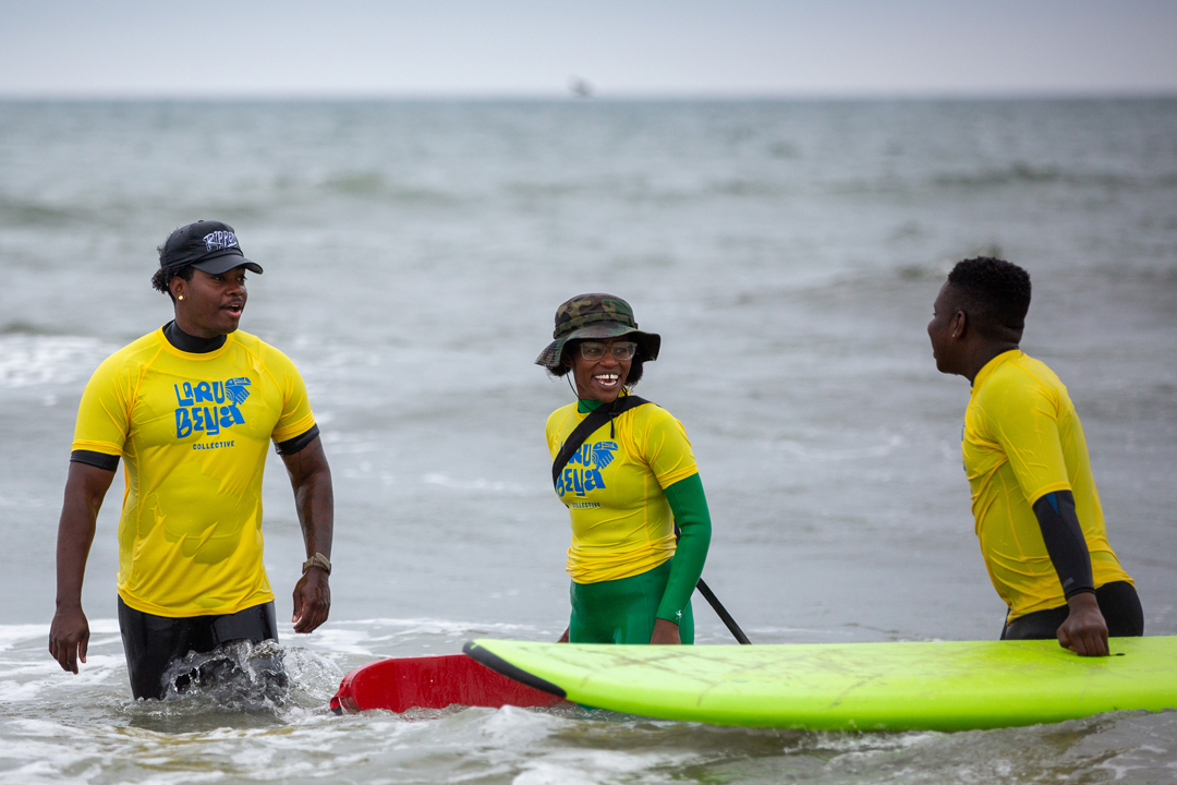 Photo of three surfers standing in the water together. A young boy holds on to his board while talking to a girl holding a life preserver and another man standing in the water.