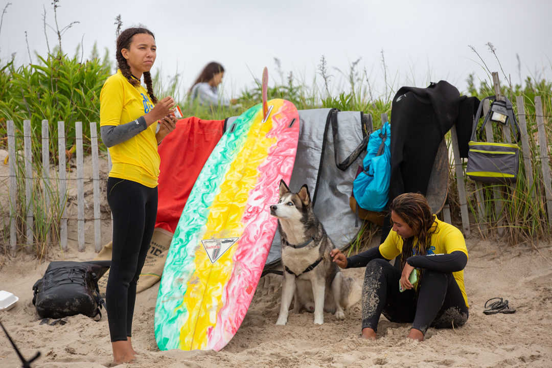 Two girls on the beach setting with surf gear. One stands next to the multicolored surfboard looking out to the waves and another sits on the sand petting a husky dog.