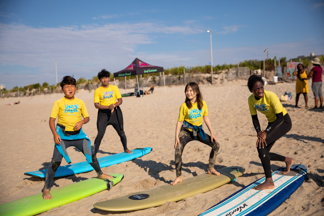 A group of kids on the beach standing on their surfboards practicing how to balance on the. They stand in squat positions to get the hang on the balance.