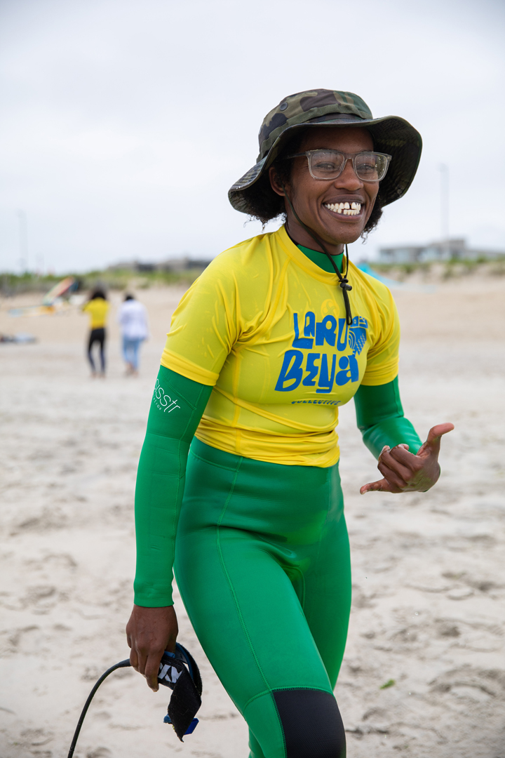 One of the surf instructors smiles for a photo as she holds the ankle leash from a surfboard in one hand and throws up a Shaka hand sign (also known as 'hang loose', that has the thumb and pinky finger extended and the three middle curled)
