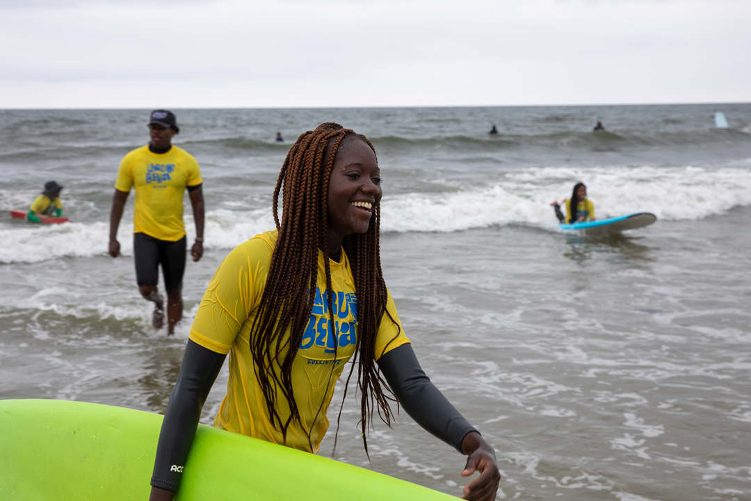 A young girl walks out of the water smiling and carrying her green surfboard. In the background there are other young surfers in the water.