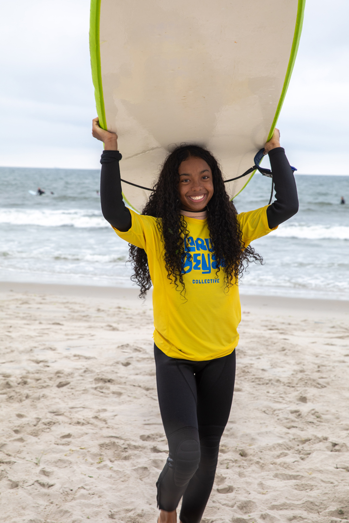 A young girl walks on the beach away from the water carrying her surfboard on her head and smiles for a picture.