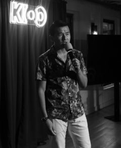 Black and white photo of Ronny Chieng performing on stage standing in front of the crowd.