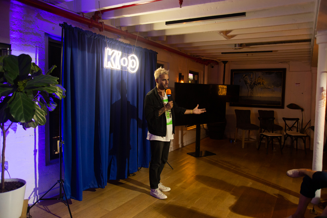 Photo of Marcus Russell Price talking on stage in front of a blue curtain and a neon sign reading KIOO.