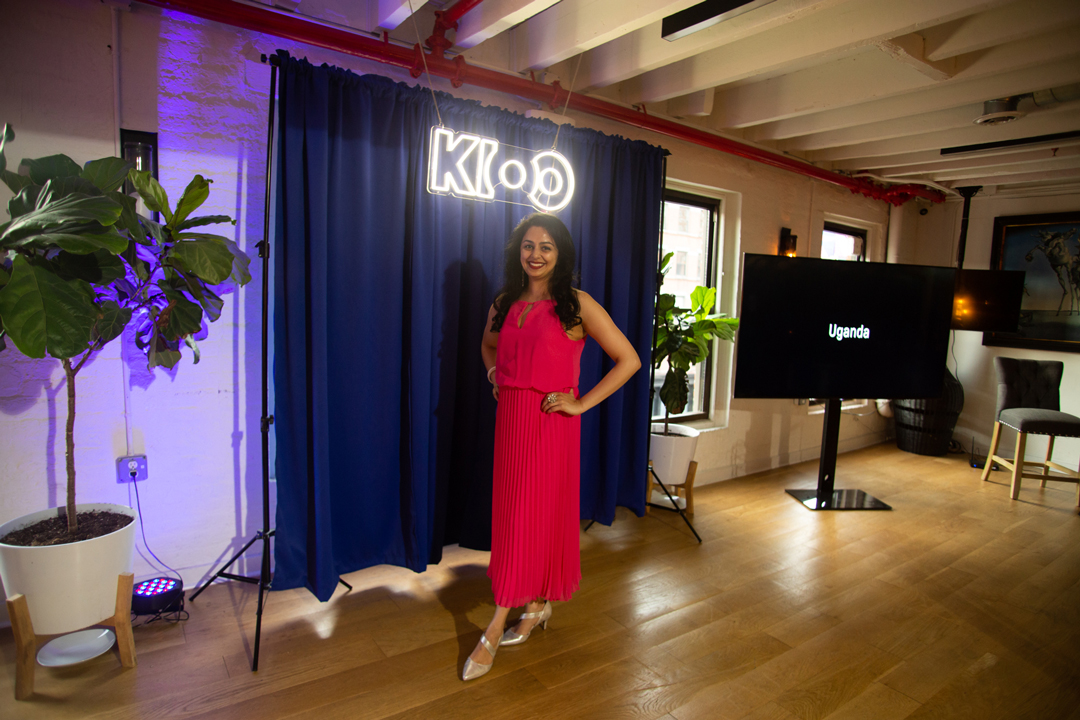 Photo of KIOO founder Babita Patel posing for a picture in front of a blue curtain and a neon sign reading KIOO.