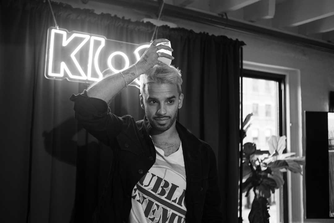Black and white photo of event host Marcus Russell Price standing in front of the stage curtain with a neon sign hanging on it reading KIOO.
