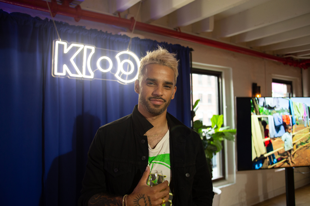 Photo of Marcus Russell Price standing in front of the blue stage curtain with a neon sign hanging on it reading KIOO.