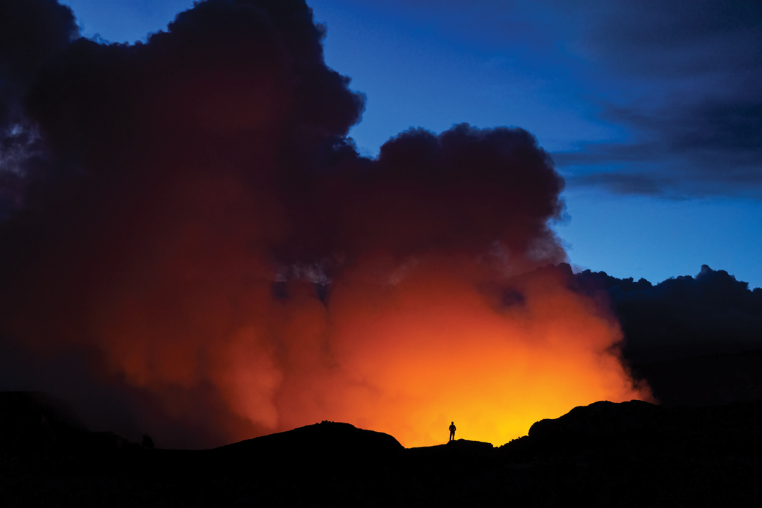One person standing at the edge of an erupting volcano in Hawaii Volcanoes National Park. The person is minuscule in size compared to the mountain of rock they stand on and the erupting smoke. The sky is the color of vivid blue in contrast to the bright orange fire from below.