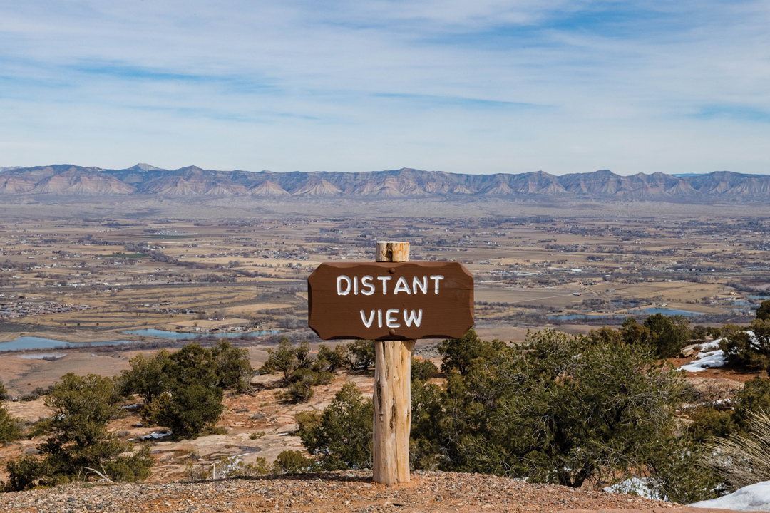 Photo of a simple wooden sign that reads "distant view". The background view stretches for miles and shows tiny houses scattered around and the Rocky Mountain range.