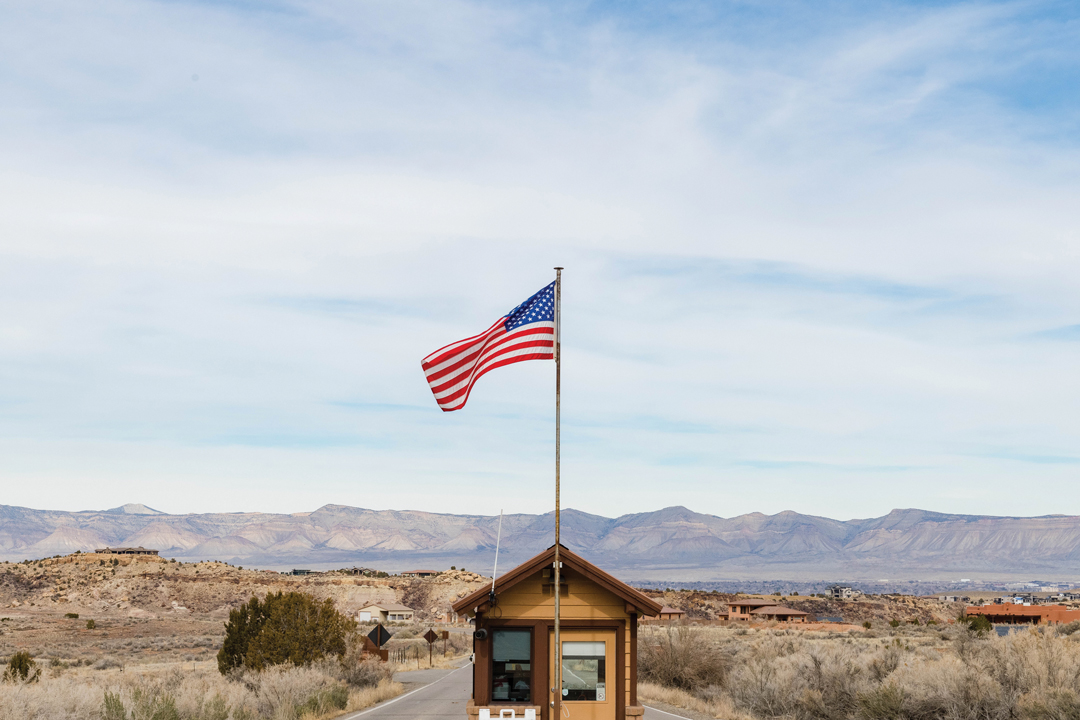 Photo of the Colorado National Monument entrance booth that has an American flag rising from it. In the background there are numerous buildings and the Rocky Mountain range.