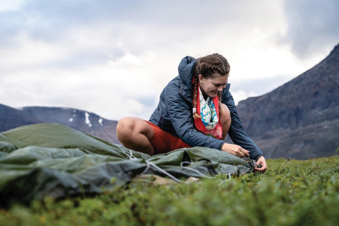 Photo of Evelina Nordin, User Engagement Specialist for Fjällräven, on a grassy hill tying up a tarp. She's surrounded by a rocky mountain range.