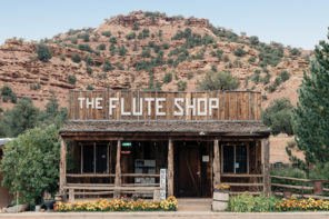 Photo of a road side stop, The Flute Shop. The small one room building has wood paneling and a short railing made from logs. Behind it is a desert mountain covered in clay-colored rocks and shrubbery.