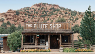 Photo of a road side stop, The Flute Shop. The small one room building has wood paneling and a short railing made from logs. Behind it is a desert mountain covered in clay-colored rocks and shrubbery.