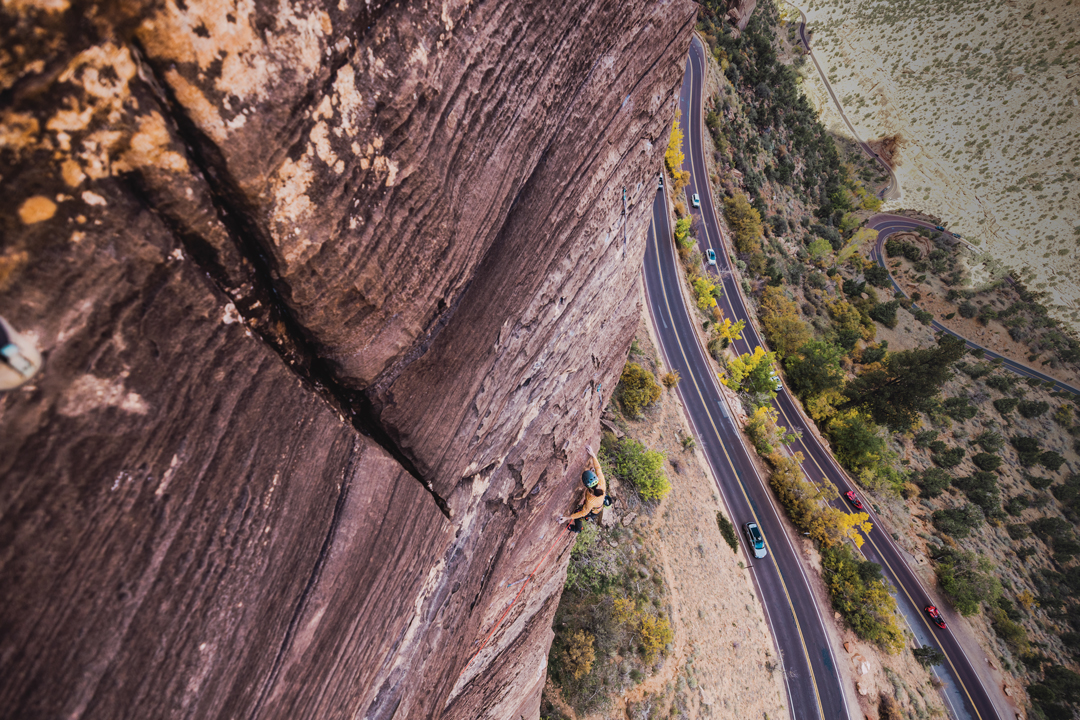 View from above of a rock climber scaling the side of a tall, flat mountain in Zion National Park. Below them, cars race by on a winding highway and the landscape is full of bushes and trees.
