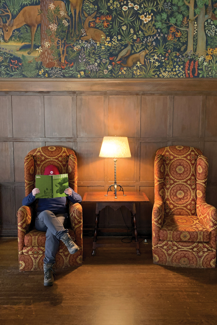 Photo of two ornate chairs against a wood paneled wall with illustrated wall paper above it. Someone is sitting in one of the chair with Whalebone's National Parks magazine covering their face.