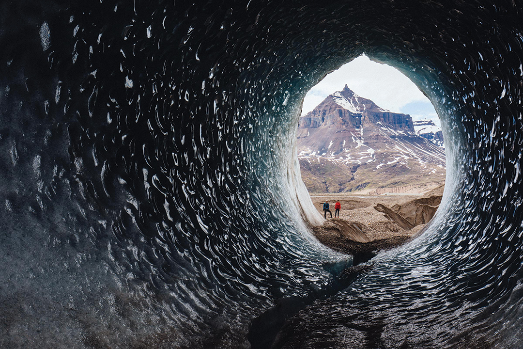 Two people staring into a large ice tube in Wrangell-St. Elias National Park and Preserve in Alaska. Water rushing through the glacier has formed it into a large circular tunnel.