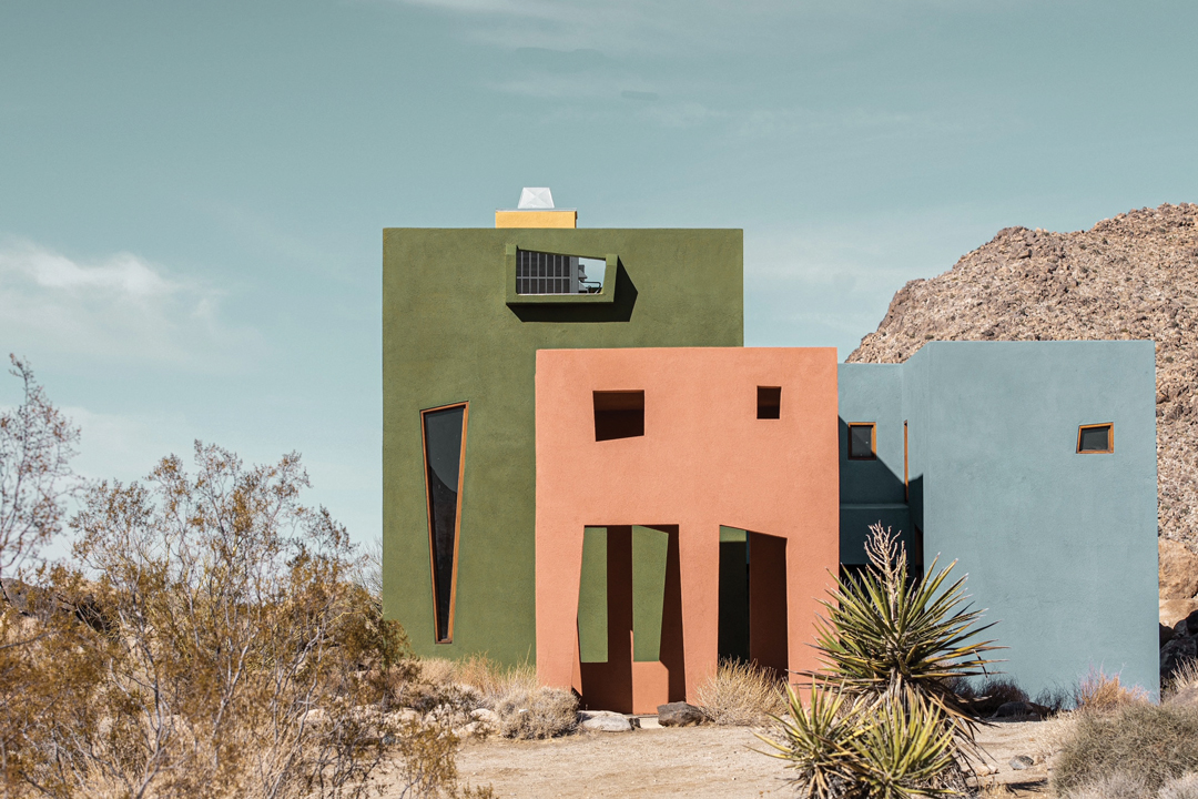 Photo of a colorful, abstract building in Joshua Tree National Park. The building is green, pink, and blue to match the color of nature around it and have abstract windows cut out from them.