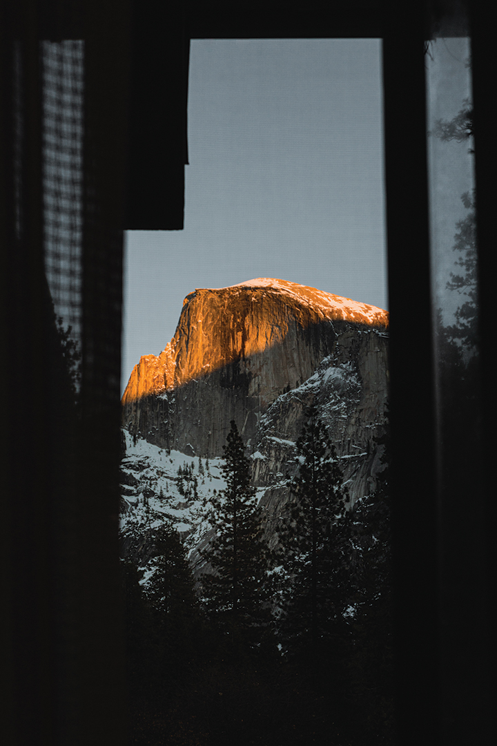 Photo of the large mountainscape in Yosemite National Park. The mountain is dusted in snow and dotted with trees. This image is taken from a windowsill so you can see the open windowpanes on the side of the image.