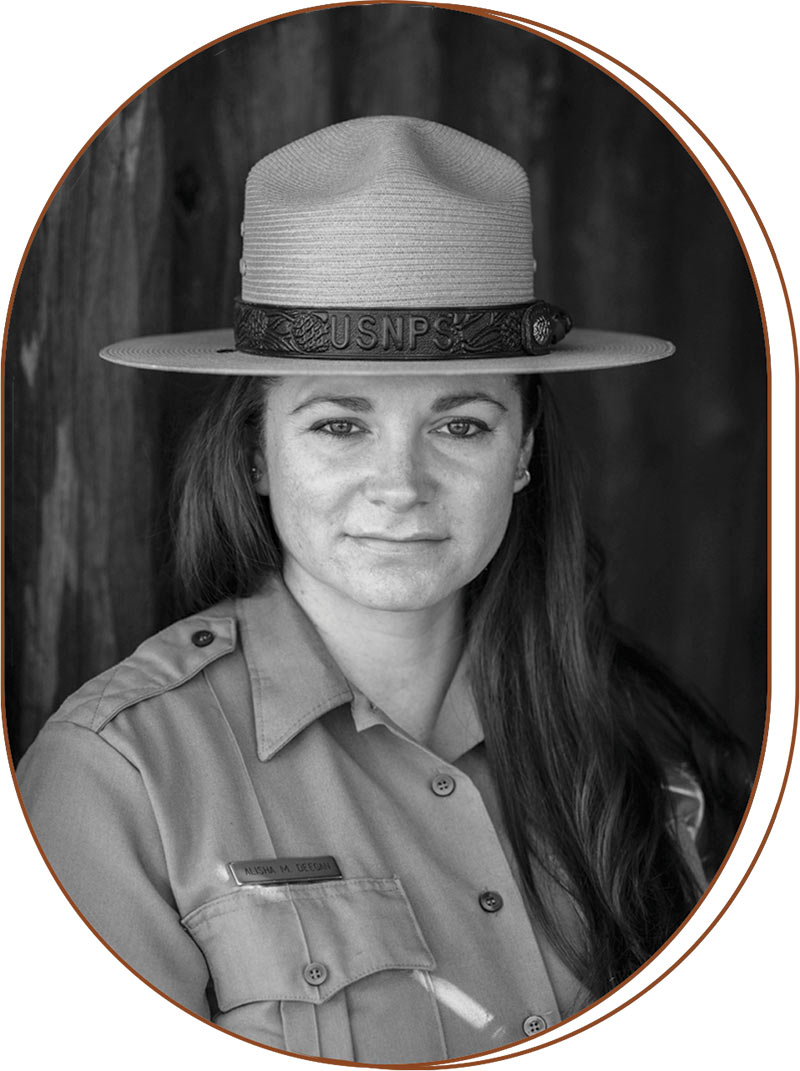 Black and white photograph of Alisha Deegan. She looks into the camera and is wearing a flat brim hate and uniform of the national park service. She has long dark hair.