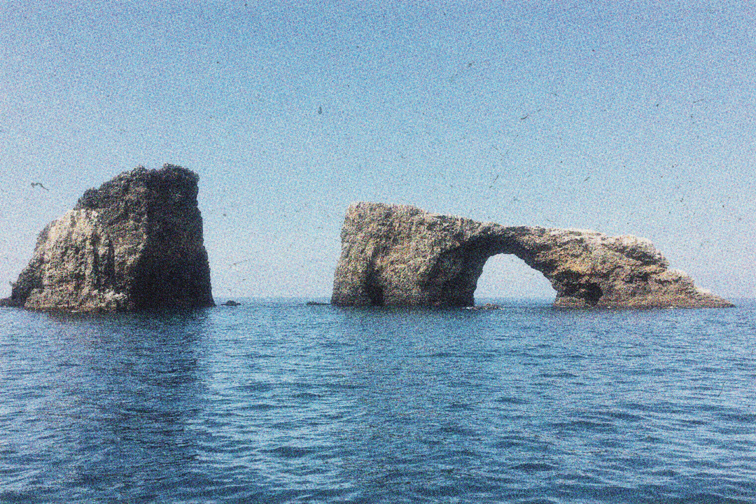 Two large rock formations halfway submerged in the ocean. One is formed into a triangle shape while the other is more rectangular and has a hole through the middle of it.