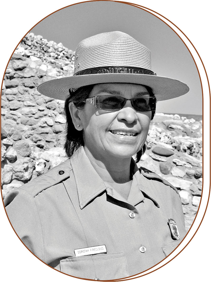 Black and white photograph of Dorothy Firecloud. She wears sunglasses, a round flat brim hat and the uniform of a national park ranger. Behind her are structures of stone and a clear sky.