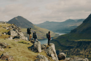 Photo of three hikers wearing backpacking gear standing on a pile of rocks overlooking a green mountain range.