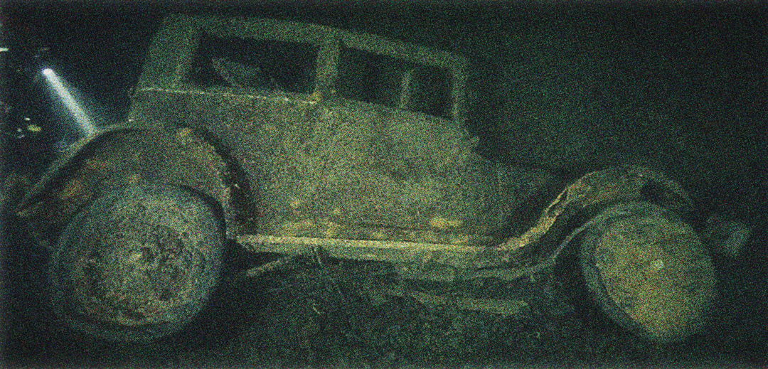 Sunken, old-fashioned car covered in rust, grime, and other buildups that's under the water.