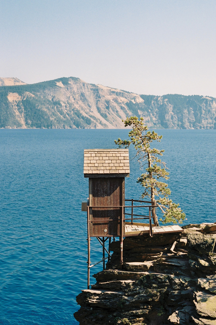 Photo of a tiny stand-alone building structure that jets off over Crater Lake. This tiny wooden building stands in front of a giant lake and mountain range.