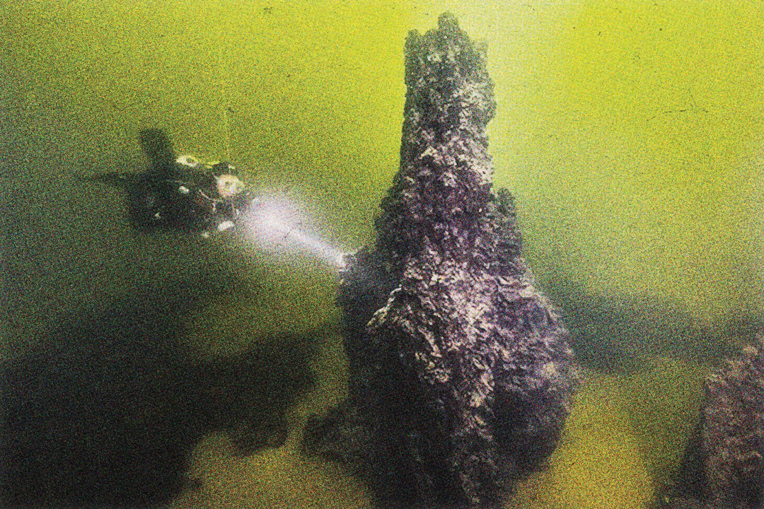 A scuba diver is investigating a large coral-like buildup under the water. The formation is significantly lager than the diver.