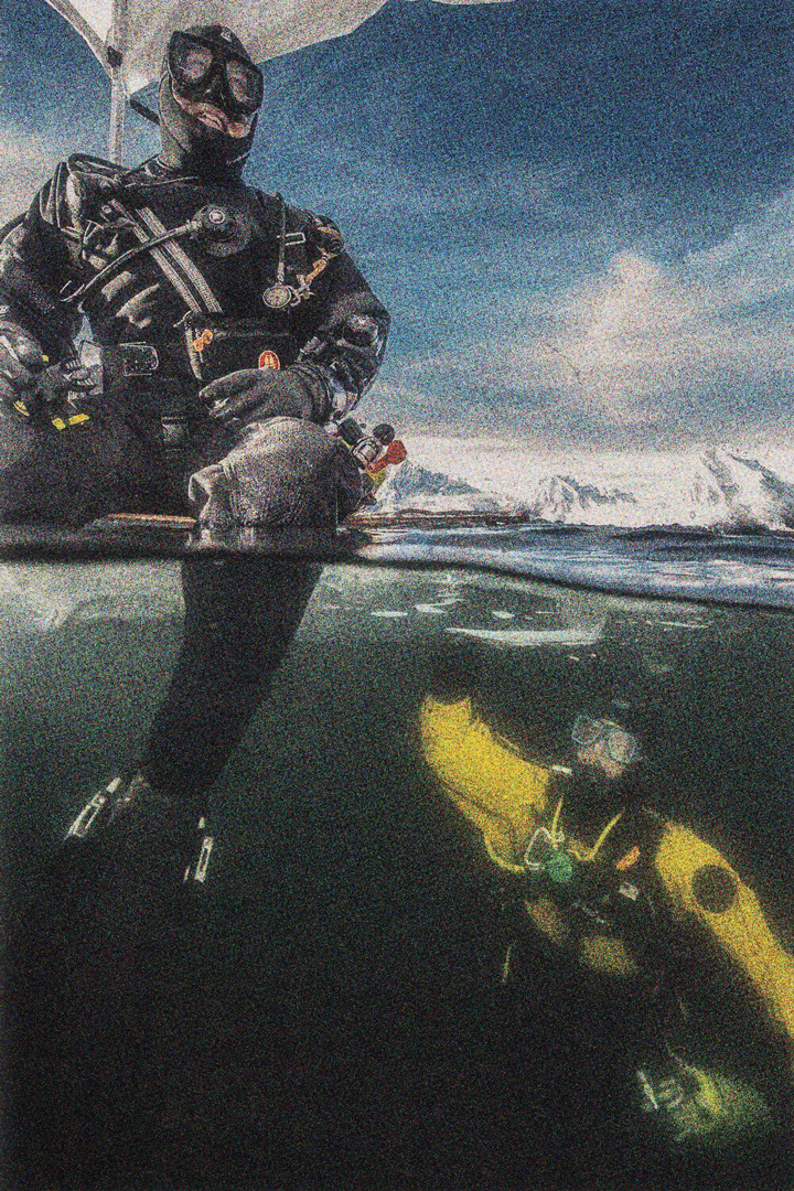 Two scuba divers in the ocean together. One sits on top of something floating in the water while you can see the other underwater reaching up towards them. In the background there is a snowy mountain range.