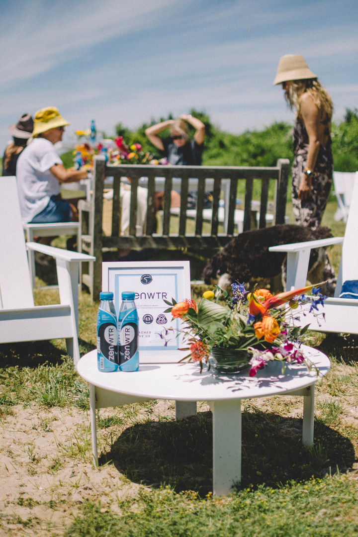 Photo of a small circular table in a yard covered with a flower arrangement, a picture frame, and bottled water. Women in the background sit at a table .