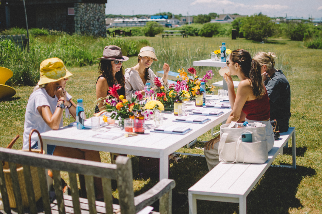Photo of five women sitting at a large table in a yard. The table is filled with flower arrangements, magazines. notebooks, and drinks.