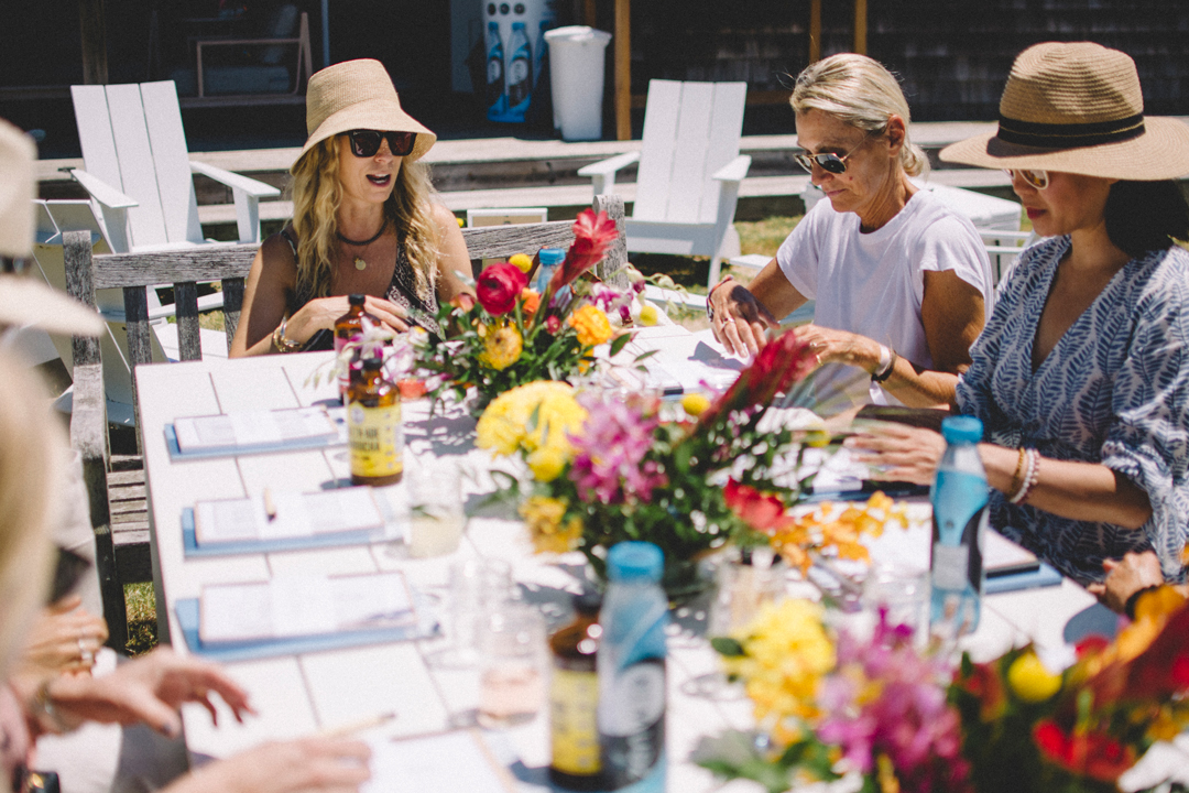 Photo of a group of women journaling at a table in a yard. The table is filled with flower arrangements, magazines, notebooks, and drinks. 