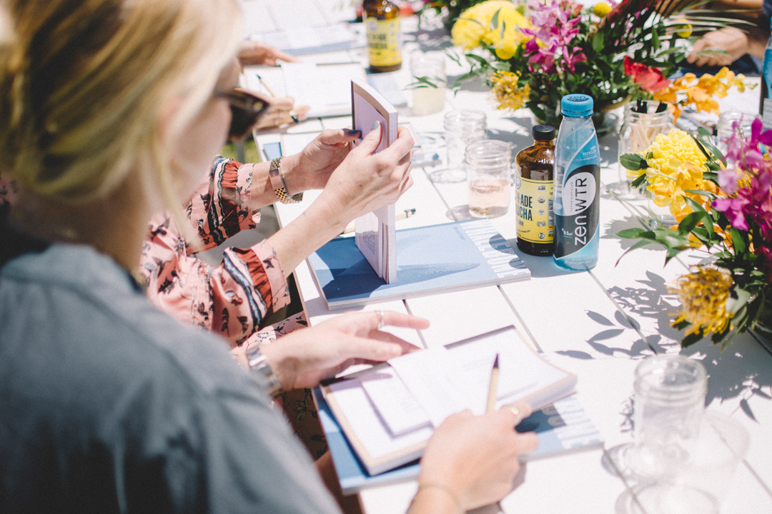 Photo of a table setting filled with flower arrangements, magazines, notebooks, and drinks. Women sit at the table holding the notebooks and papers.