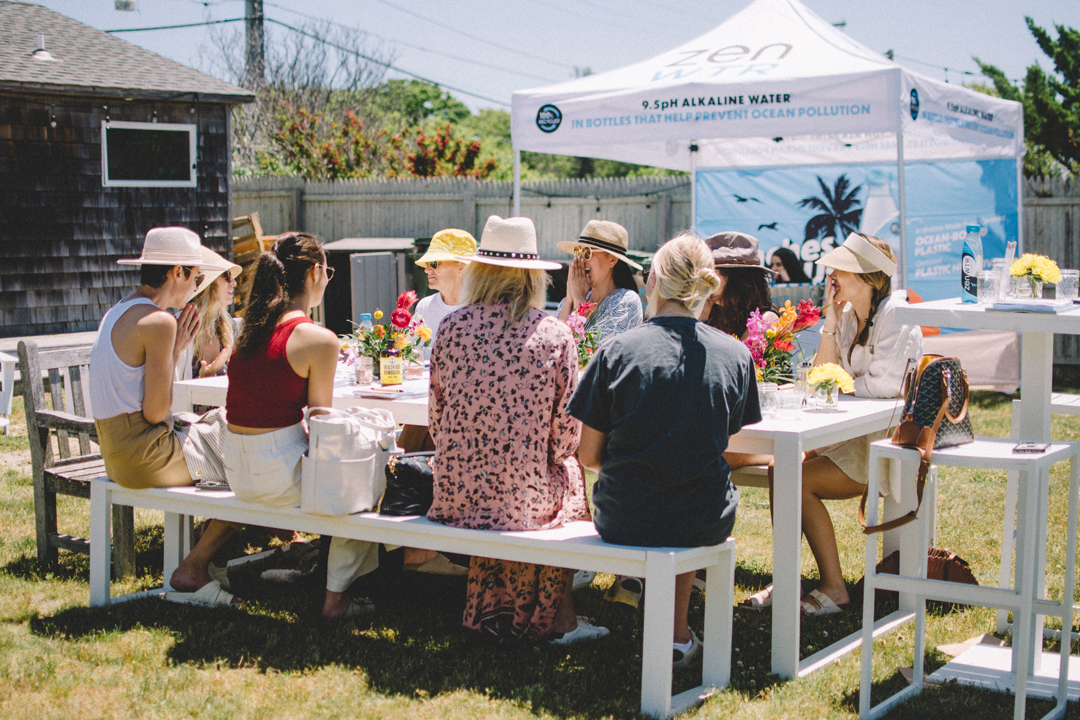 Photo of a group of women talking at a table in a yard. The table is filled with flower arrangements, magazines, notebooks, and drinks. There is a tent behind them handing out water.