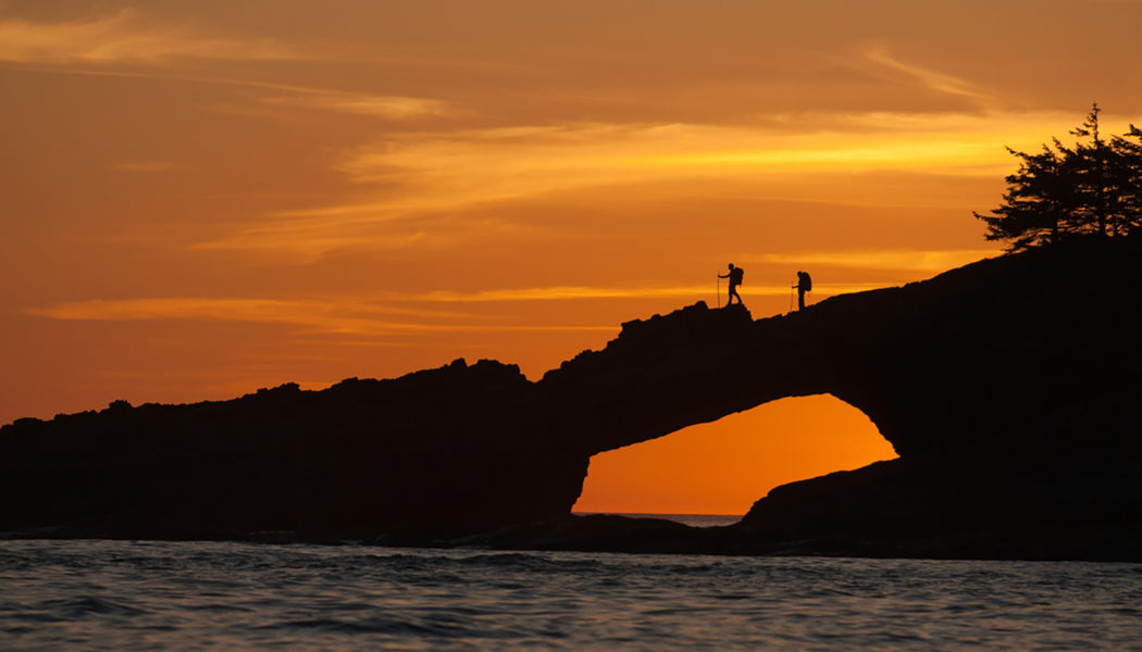 Photo of two hikers silhouetted against an orange sky as they walk over a rocky pass that goes over the ocean.