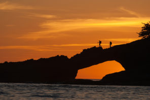 Photo of two hikers silhouetted against an orange sky as they walk over a rocky pass that goes over the ocean.