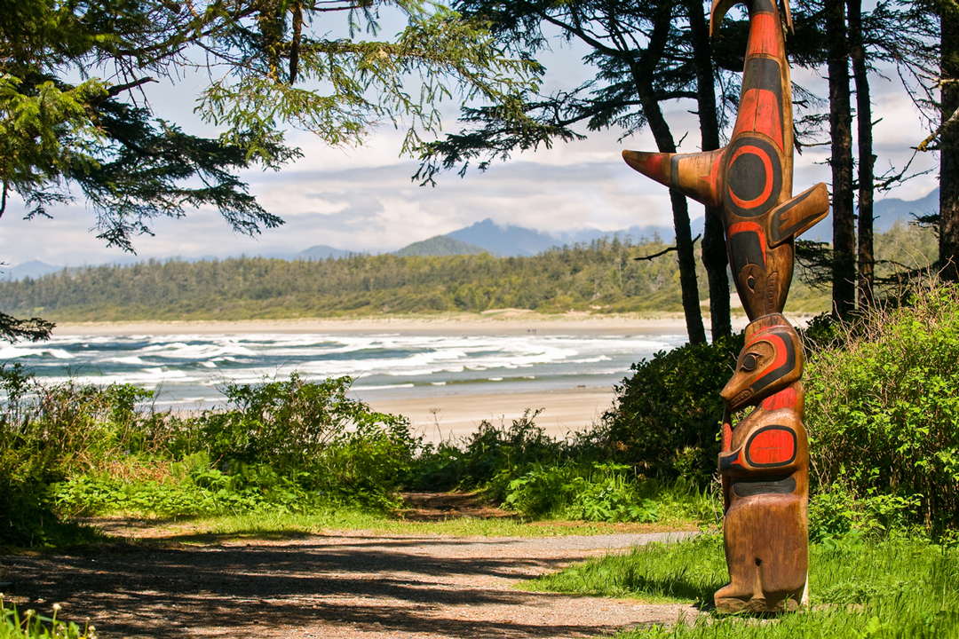 Photo of an orange and black totem poll on the side of a trail in the woods. In the background is a large forest waves crashing onshore to the beach.