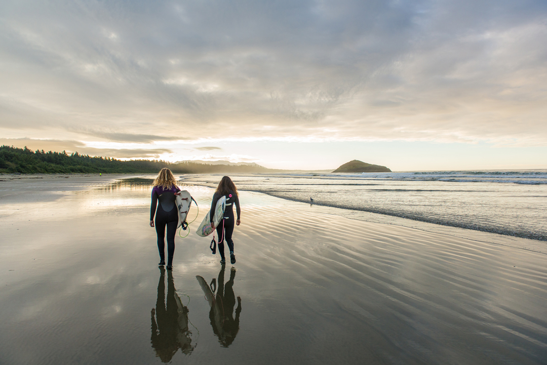 Photo of two surfers walking along the beach  at sunrise carrying their boards. In front of them is a vast forest and a mound of rocks offshore.